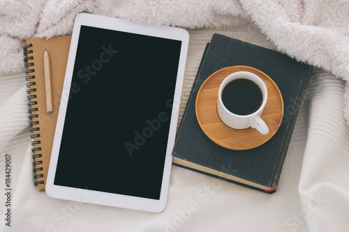 Cup of coffee on the old book and tablet device with empty black screen over cozy and white blanket. Top view. photo