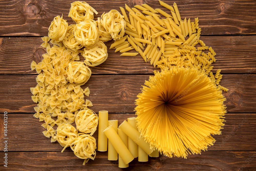 Different kinds of pasta on a wooden background. Farfalle, fettuccine, noodles, fusilli and penne rigate. Tasty Italian cuisine. Different kinds of pasta on a wooden background. Farfalle, fettuccine, noodles, fusilli and penne rigate. Tasty Italian cuisine.