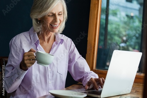 Smiling senior woman drinking coffee while working on laptop photo