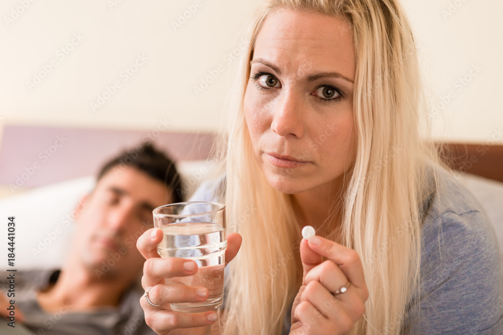 Young worried woman sitting in bed next to her partner while taking a pill before sleep at night Young worried woman sitting in bed next to her partner while taking a pill before sleep at night