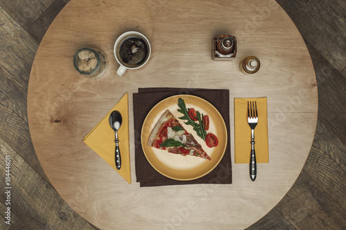 flat lay with arranged piece of pizza on plate, cup of tea and cutlery on wooden tabletop