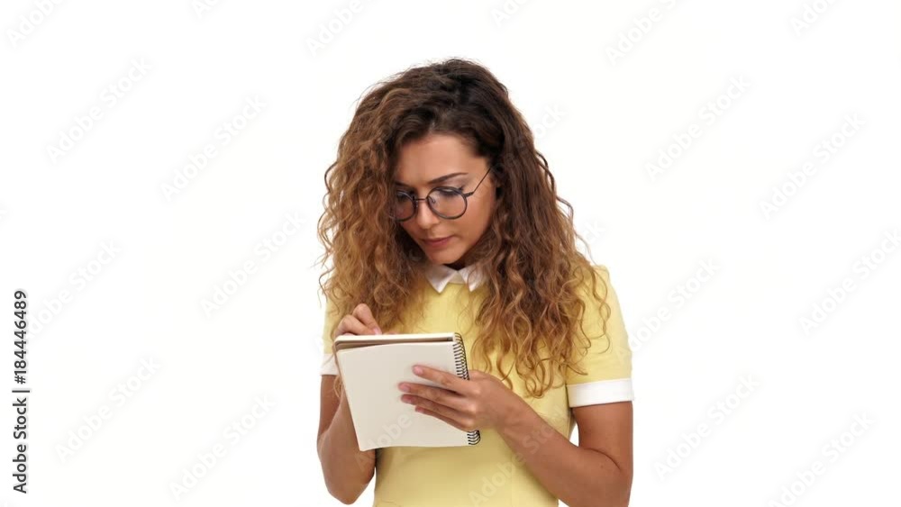 Young Concentrated curly brunette woman in dress and eyeglasses listening and writing something in notebook over white background Young Concentrated curly brunette woman in dress and eyeglasses listening and writing something in notebook over white background