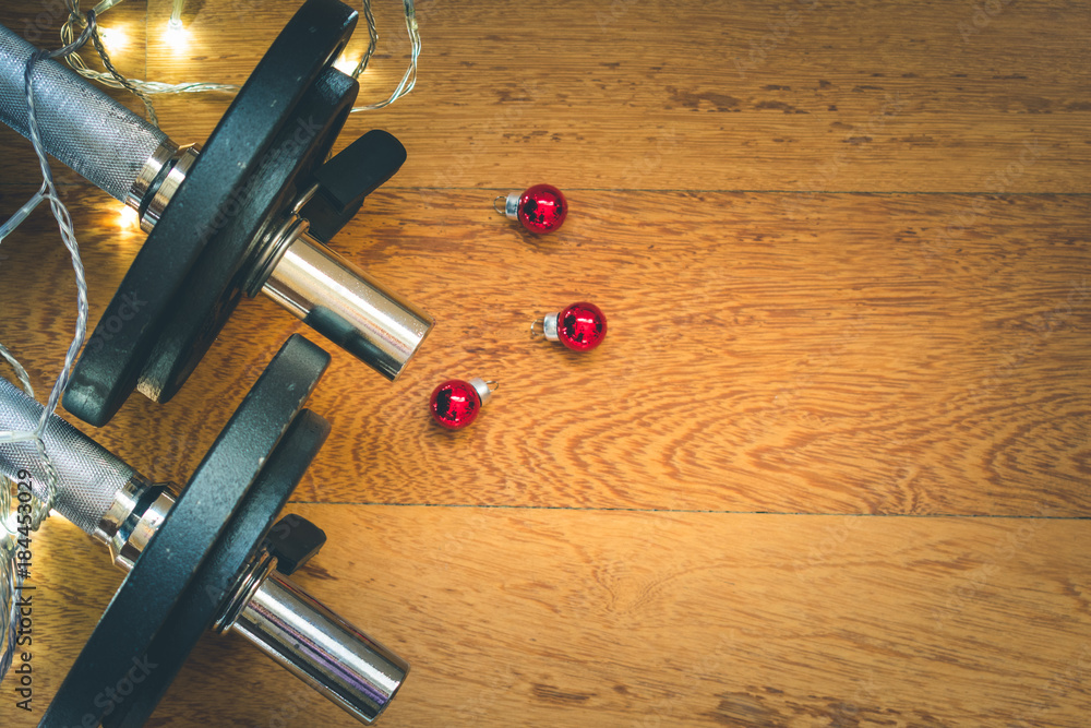 Top view of black metal fitness weights on wooden background with shining Christmas lights and red decorations, holidays and fitness concept