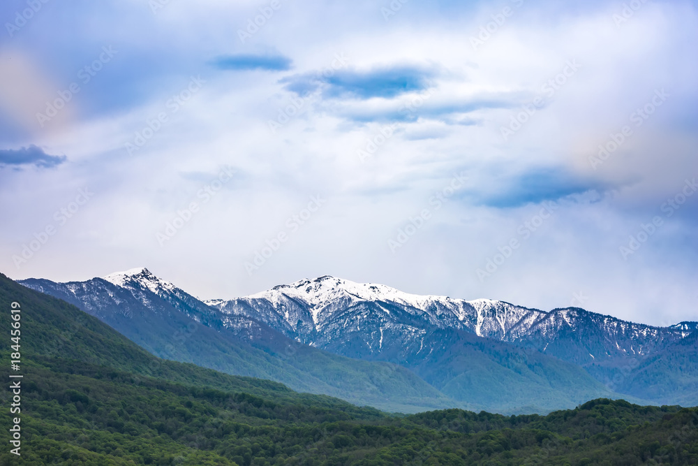 Fototapeta premium The mountain range of the Caucasus with a snowy peak in the spring with a bright green forest. Mountain valley on the way to Krasnaya Polyana from Adler, Sochi, Russia.