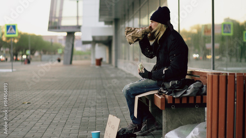 Homeless young man eating sandwich and drinking alcohol from paper bag on bench at city street in evening © silverkblack
