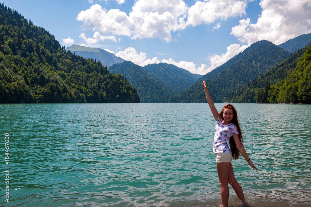 A smiling girl with long hair and open arms against the backdrop of the high mountain lake Ritsa in Abkhazia.