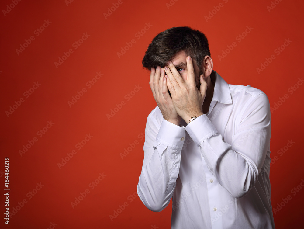 Unhappy thinking grimacing bearded business man covering the face his hands in white shirt and looking on bright orange background with empty space background. Unhappy thinking grimacing bearded business man covering the face his hands in white shirt and looking on bright orange background with empty space background.