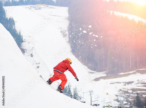 Young male snowboarder riding from the top of the snowy hill with snowboard in the evening at sunset at ski resort. Ski season and winter sports concept