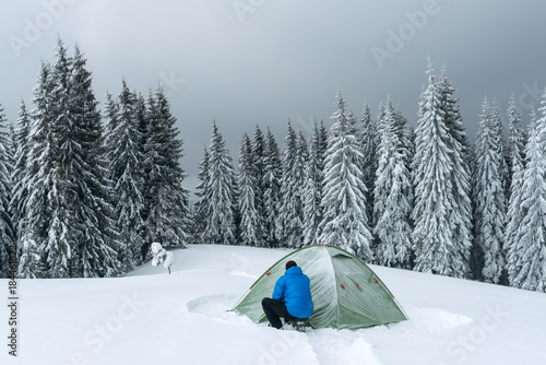 Green tent and tourist against the backdrop of snowy pine tree forest. Amazing winter landscape. Tourists camp in high mountains. Travel concept