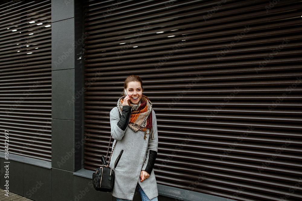 Fototapeta premium Street style. The girl communicates on the smartphone. The woman is dressed in jeans and a gray jacket, she costs on the street in the city against the background of an impressive gray wall.