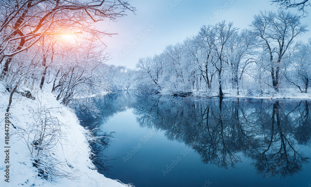 Winter forest on the river at sunset. Colorful landscape with snowy trees,  frozen river with reflection