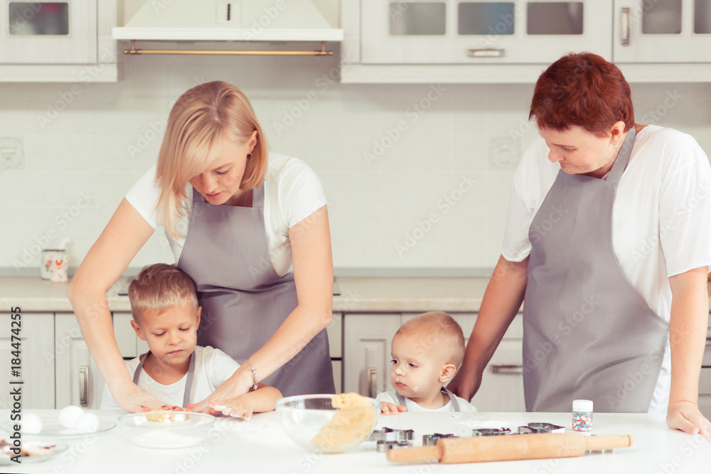 Cooking gingerbread cookies at home in the white kitchen. Happy family of mother, grandmother and little kids boys in the kitchen. Mom, grandma and children sons cooking together Cooking gingerbread cookies at home in the white kitchen. Happy family of mother, grandmother and little kids boys in the kitchen. Mom, grandma and children sons cooking together