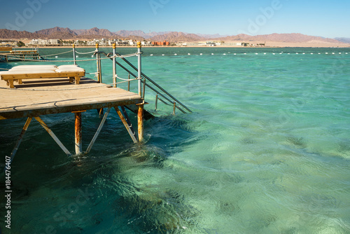 Pier with chaise longues in the sea in resort. Summer vacation. View at a clear sea with turquoise water. Summer vacation at a sea coastline in an exotic country. Staircase to the water for swimming.