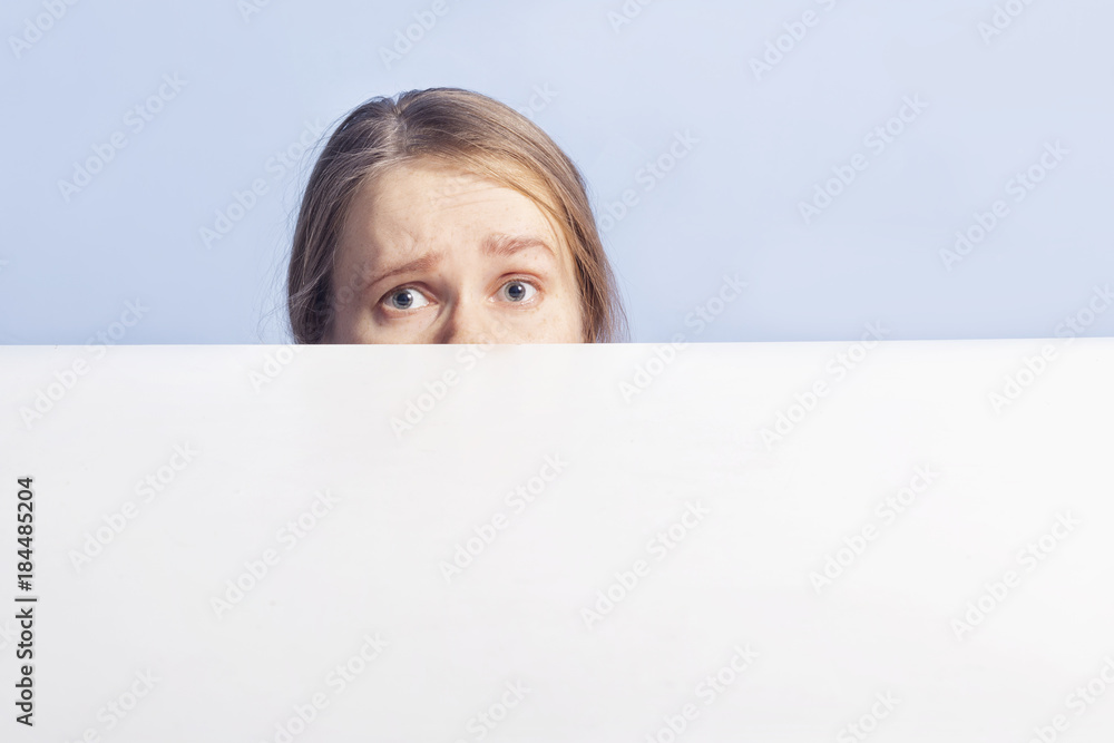 girl looks confused behind white banner, isolated on blue background girl looks confused behind white banner, isolated on blue background