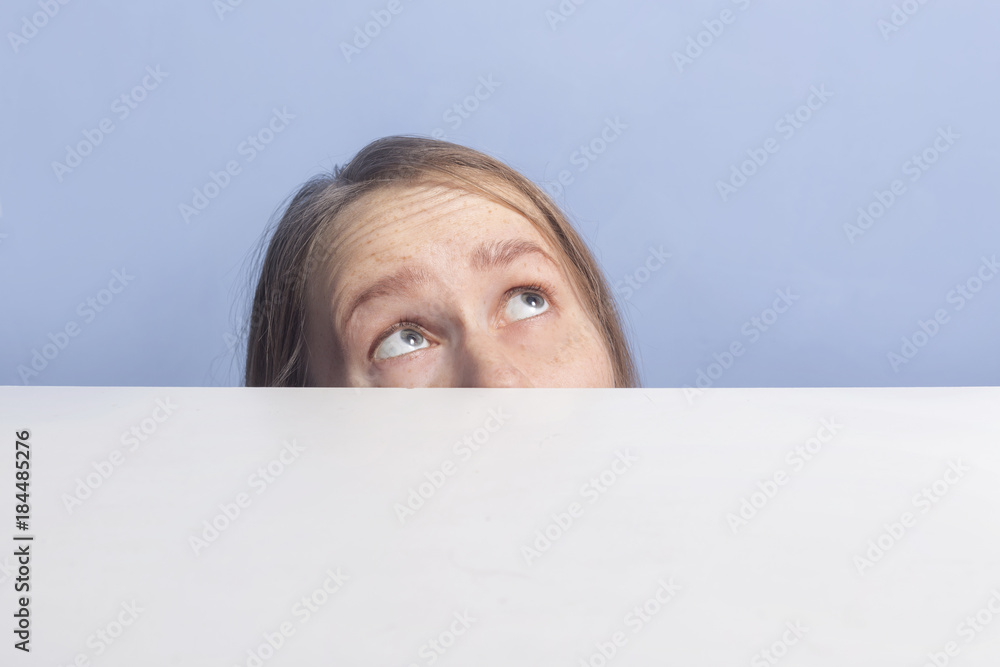 brown-haired girl looks up in surprise behind white banner, isolated on blue background brown-haired girl looks up in surprise behind white banner, isolated on blue background