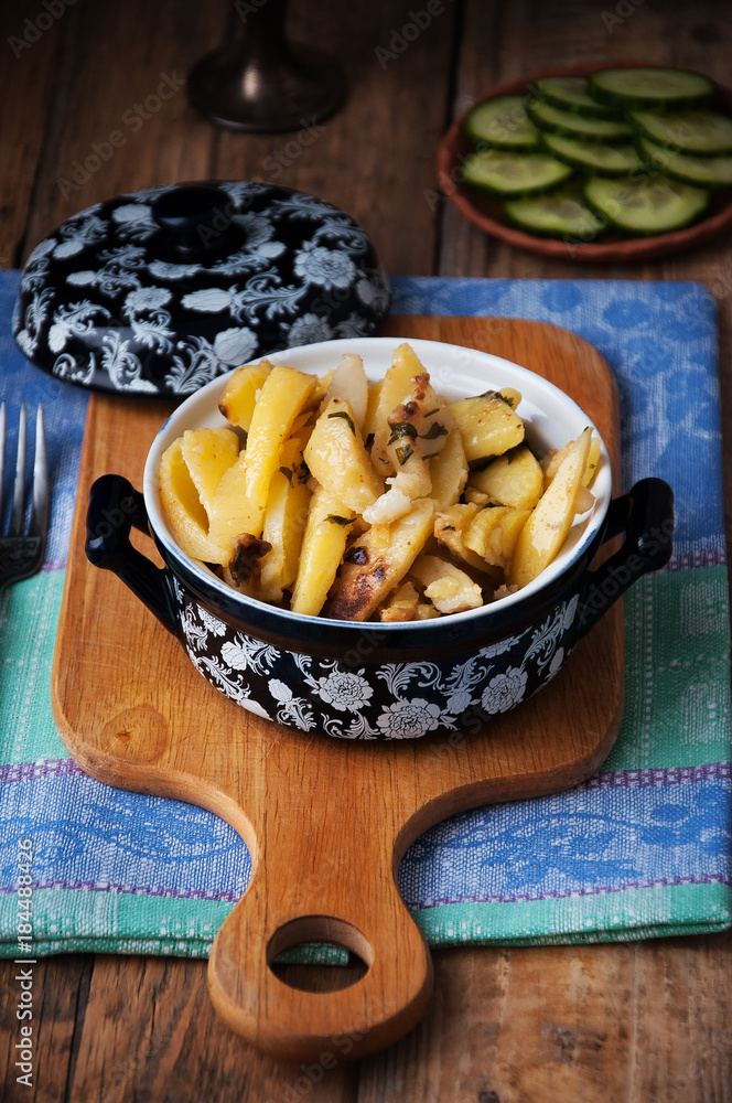On a wooden table a dish of fried potatoes, in the background a plate with fresh cucumbers, sliced ​​slices