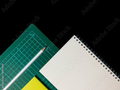 Clear color note book opened for note or lecture note or memo for remine and green cutting mat with black head color pencil in white apparel and orange posit on black background.