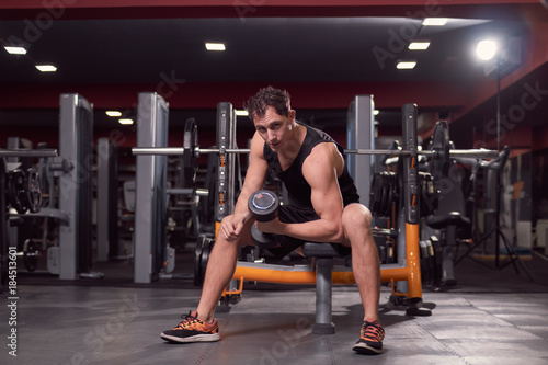 one young man, dark gym indoors fitness equipment, one hand dumbbell biceps exercise, sitting bench, looking at camera.