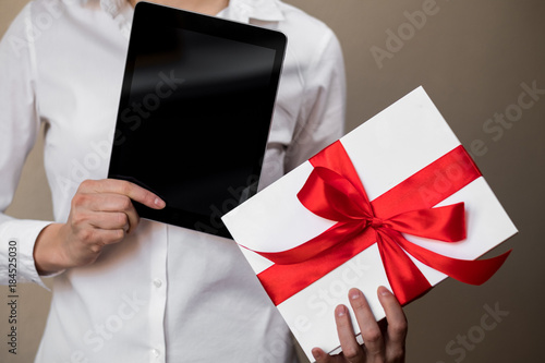 A woman in a white shirt is holding a gift, a black tablet and white box decorated with a red bow.