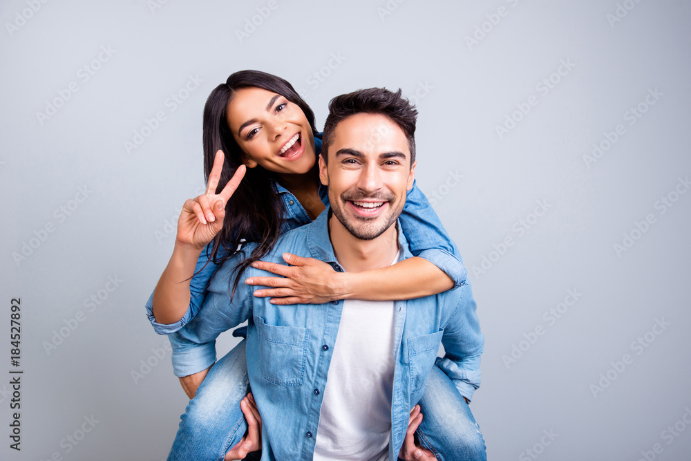 he and she travel together, love story of attractive, happy, smiling, sweet couple in casual outfit, man carrying his lover on back, woman showing peace symbol over grey background he and she travel together, love story of attractive, happy, smiling, sweet couple in casual outfit, man carrying his lover on back, woman showing peace symbol over grey background