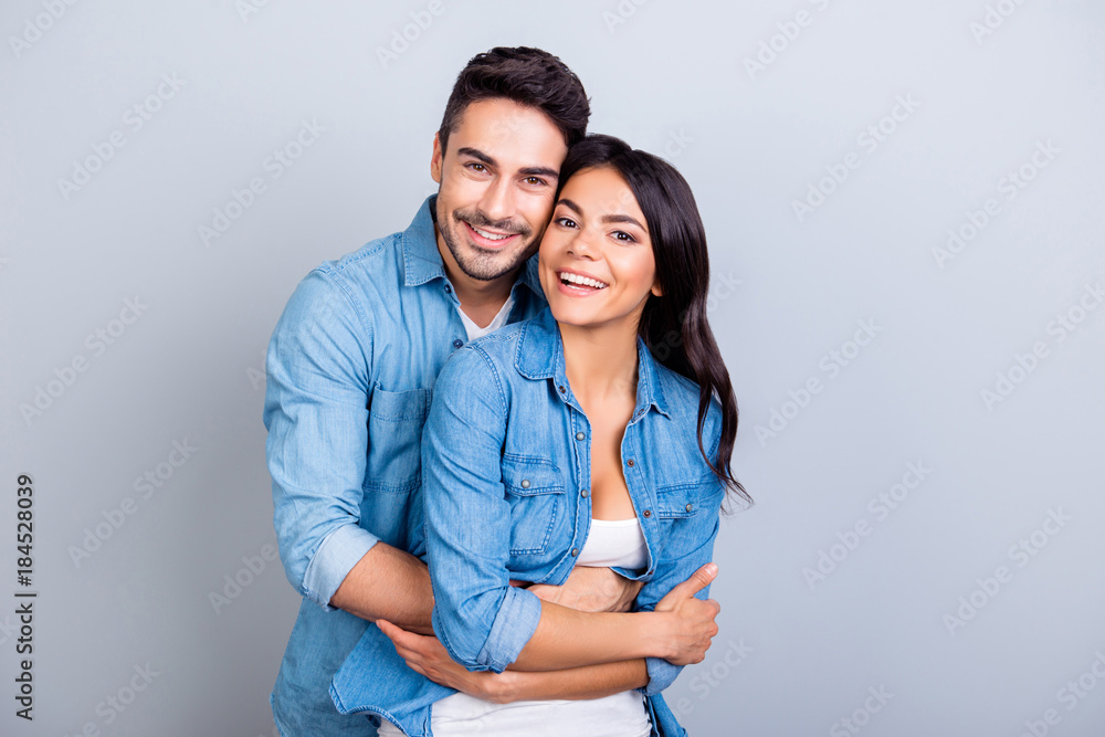 Portrait of cheerful lovely cute couple with beaming smiles hugging and looking at camera over grey background - obrazy, fototapety, plakaty 