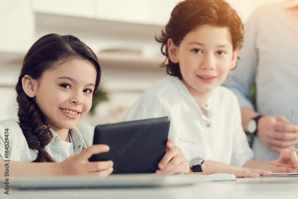 Modern device. Pleasant cheerful cute girl sitting at the table with her brother and smiling while using a tablet Modern device. Pleasant cheerful cute girl sitting at the table with her brother and smiling while using a tablet