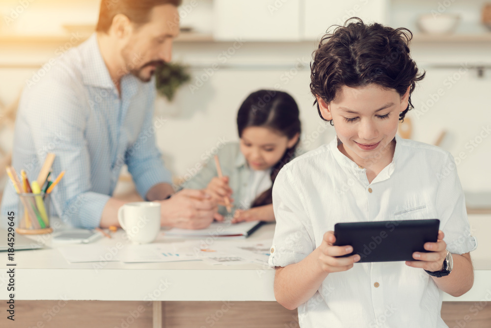 Modern igen. Nice positive smart boy smiling and looking at the tablet screen while using it for plying games Modern igen. Nice positive smart boy smiling and looking at the tablet screen while using it for plying games