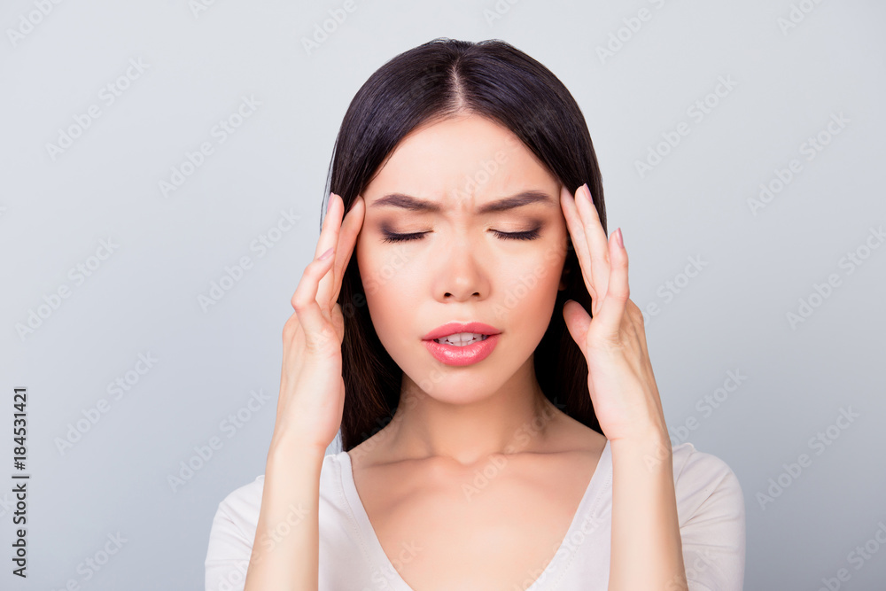 Close up portrait of confident brunette beautiful student girl having a headache, stress, she has eyes closed and she is touching her temple while standing over grey background Close up portrait of confident brunette beautiful student girl having a headache, stress, she has eyes closed and she is touching her temple while standing over grey background