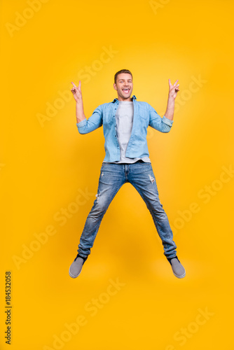 Go, mad! Glad, funny, funky guy in casual outfit full of energy is jumping up, fooling, demonstrating v-sign with two hands, isolated over yellow background