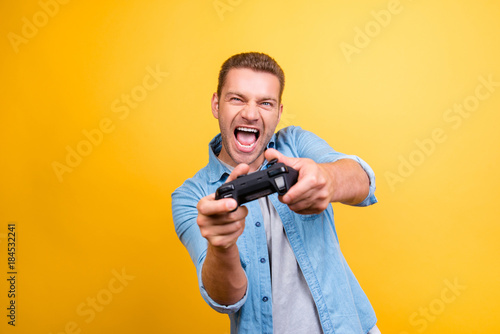 Portrait of young, cheerful, attractive, very excited guy holding joystick and playing video games over yellow background