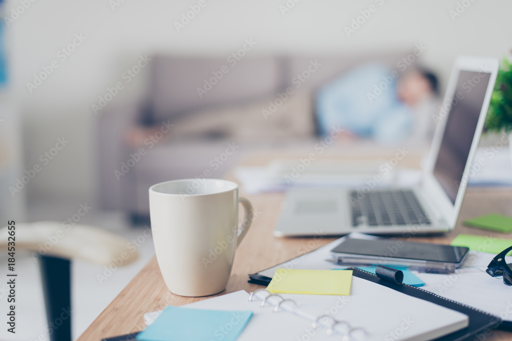 Concept of working all day and night long. Close up photo of white cup of coffee standing on desk near notes and laptop, tired depressed worker is sleeping on the background, blurred background Concept of working all day and night long. Close up photo of white cup of coffee standing on desk near notes and laptop, tired depressed worker is sleeping on the background, blurred background