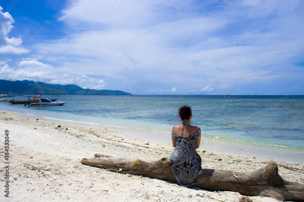 young Asian woman sitting on trunk at white sand beach in Bali Indonesia looking at the blue sea horizon under a summer blue sky young Asian woman sitting on trunk at white sand beach in Bali Indonesia looking at the blue sea horizon under a summer blue sky