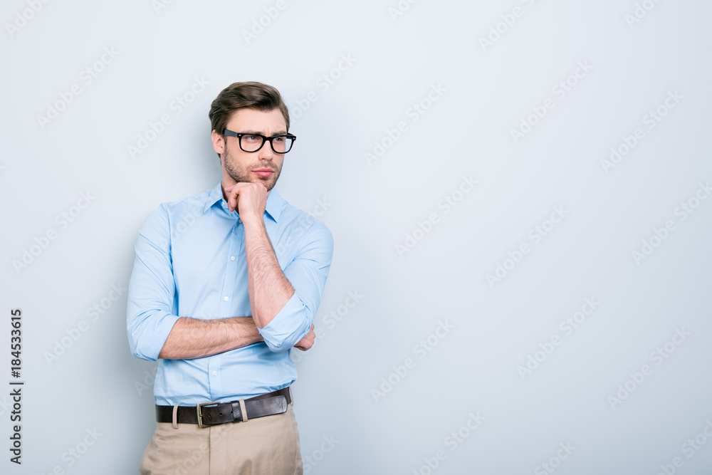 Portrait with copy space of attractive, clever, stunning man in shirt, pants and glasses touching chin with hand looking to the side with thoughtful expression over grey background Portrait with copy space of attractive, clever, stunning man in shirt, pants and glasses touching chin with hand looking to the side with thoughtful expression over grey background