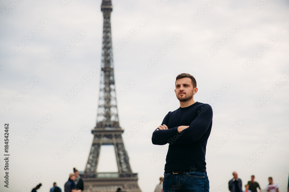 A young man wearing a dark blue jacket is standing on the background of the Eiffel Tower. Sunny weather is autumn