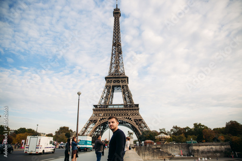 A young man wearing a dark blue jacket is standing on the background of the Eiffel Tower. Sunny weather is autumn © Aleksandr