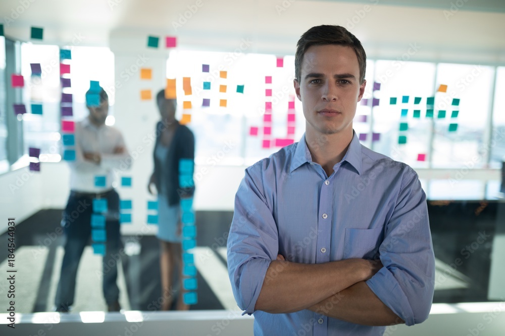 Male executive standing in office with colleagues in background Male executive standing in office with colleagues in background