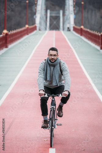 handsome adult man in sweater riding bicycle on pedestrian bridge