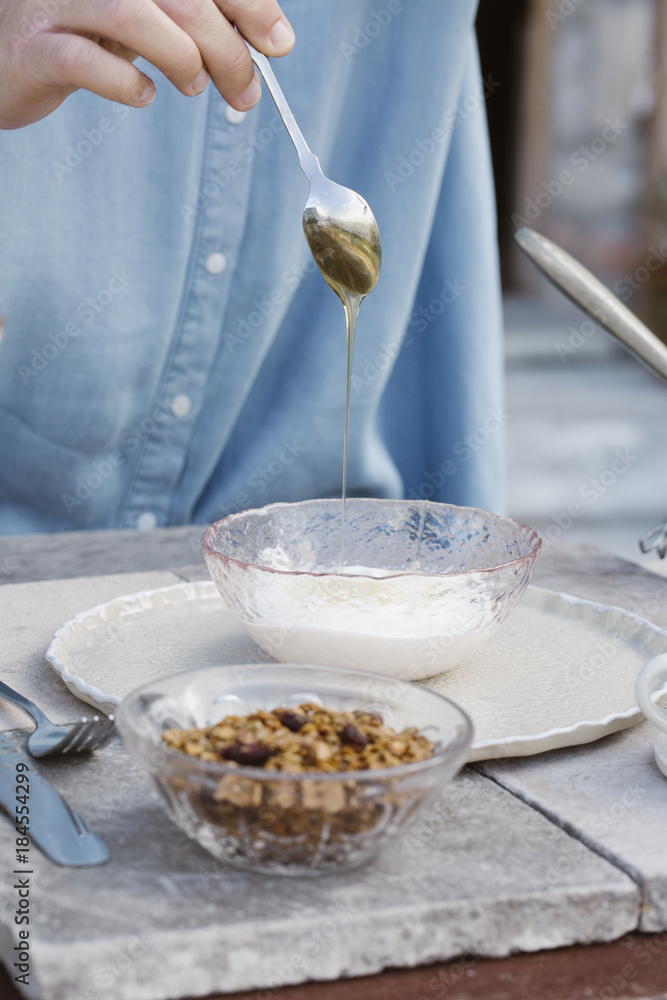 Italy, woman dripping honey on natural yoghurt at breakfast table on terrace, partial view Italy, woman dripping honey on natural yoghurt at breakfast table on terrace, partial view
