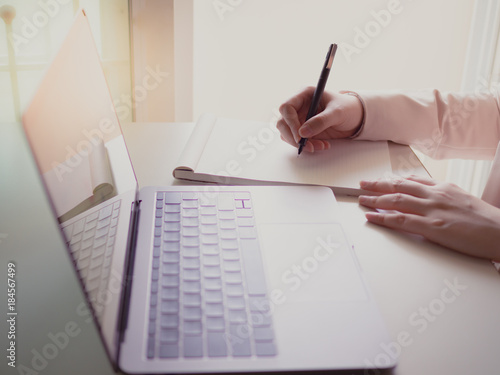 asian business woman(30s to 40s) record on blank book by black pen with pink or pastel suits with soft focus keyboard of laptop foreground