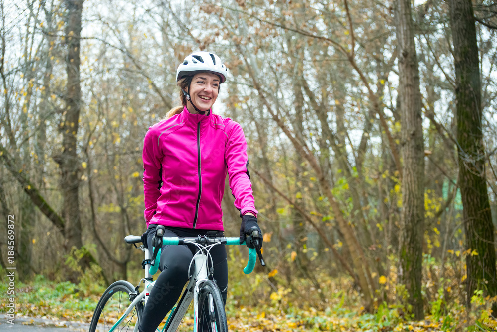 Fototapeta premium Portrait of Young Smiling Female Cyclist in Pink Jacket Resting with Road Bicycle in the Cold Sunny Autumn Day
