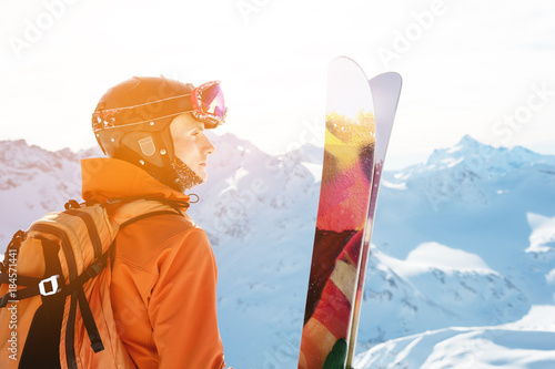 A skier in an orange overall with a backpack on his back wearing a helmet and with ski poles in his hands is standing on a precipice in front of a snowy abyss in the background of a beautiful photo