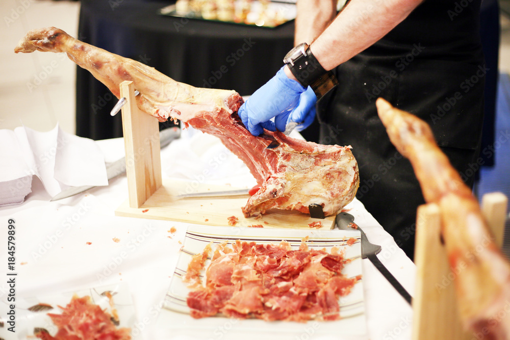 Hands of a man in gloves in the market or a store cut out a slice of the traditional Spanish jamon