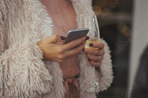 Trendy woman in pink fur coat using mobile phone while having a glass of champagne in restaurant photo