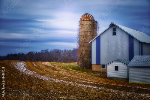 A quiet midwestern farmstead in winter, with glowing sky, silo and blue and white barn