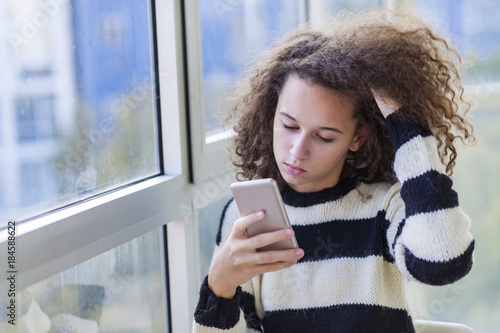 Curly hair teen girl with mobile phone by window
