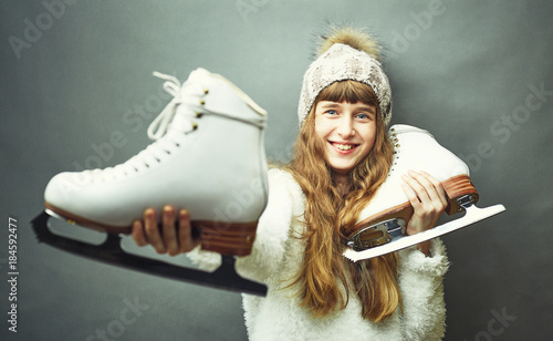 A girl with skates in her hands in a white sweater and hat.
