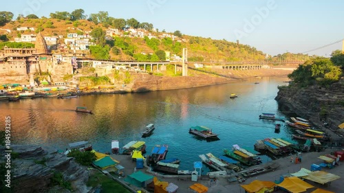 Sunset time lapse Omkareshwar cityscape, India, sacred hindu temple. Holy Narmada River, boats floating. Travel destination for tourists and pilgrims. photo