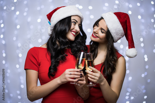 Portrait of two women in santa hats and red dress cheers and drink champagne on bokeh light background. Winter holiday Christmas and New Year concept
