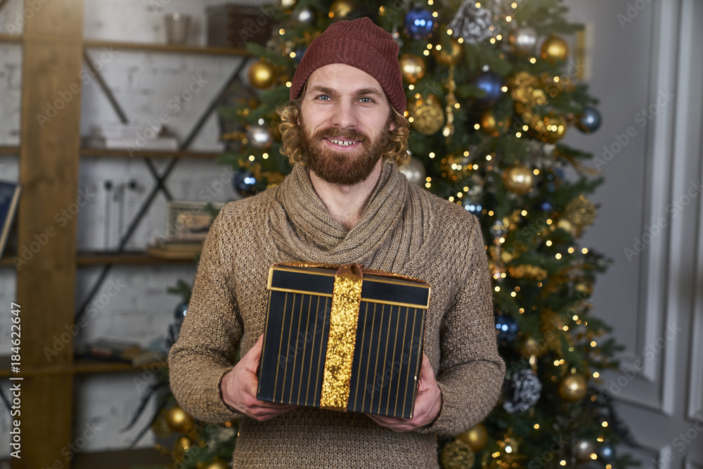 Fototapeta premium Young man cheerful in a sweater gift in a hand Christmas tree on background, joy, happiness, new year