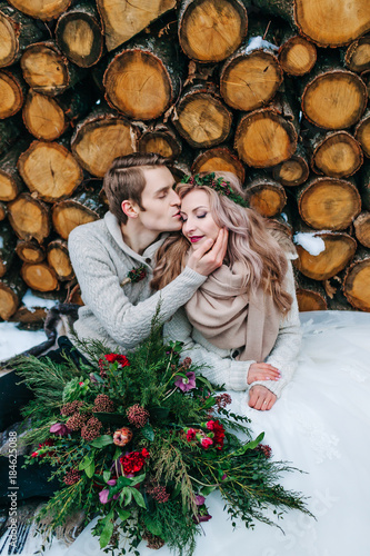 Bride and groom tenderly looks at each other. Stylish newlyweds sits on snow on the wooden background. Winter wedding photo